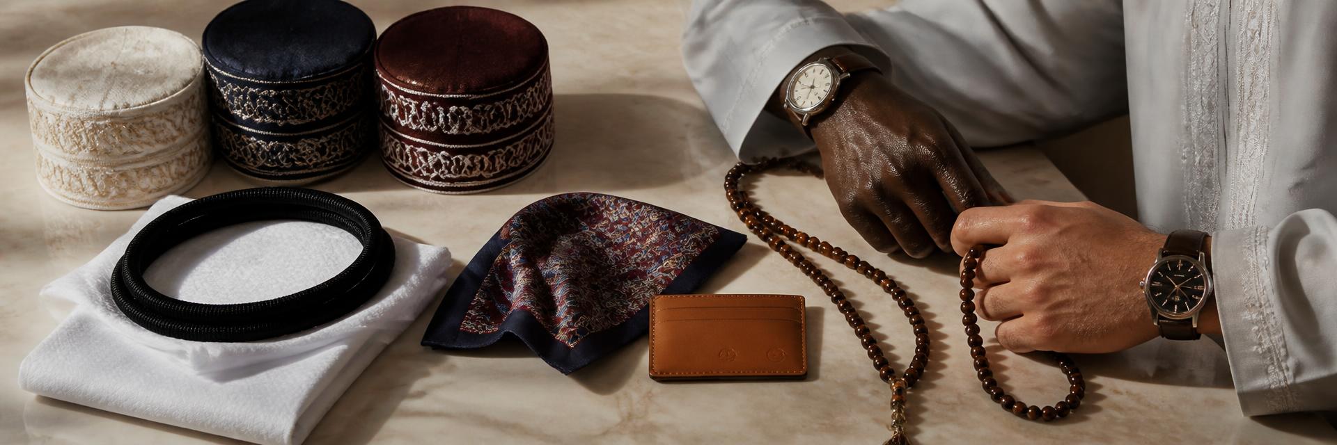 Menswear accessories flat-lay on warm cream marble — embroidered kufi caps in cream/navy/burgundy, a folded white ghutra with black agal, a leather-strap watch, dark wood and amber prayer beads (tasbih), a silk pocket square and a caramel cardholder. Two hands modeling — one Black man's hand and one Middle Eastern man's hand — with tailored thobe cuffs visible.
