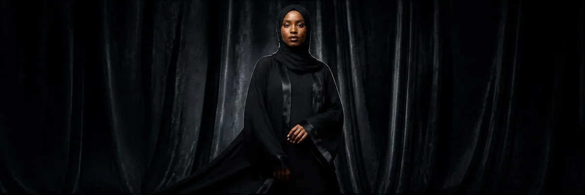 Portrait of a Black woman modeling a flowing jet-black abaya with satin trim and matching silk hijab against a dark velvet studio backdrop with dramatic rim lighting — Emaanly abaya edit. Hair, neck and body fully covered.