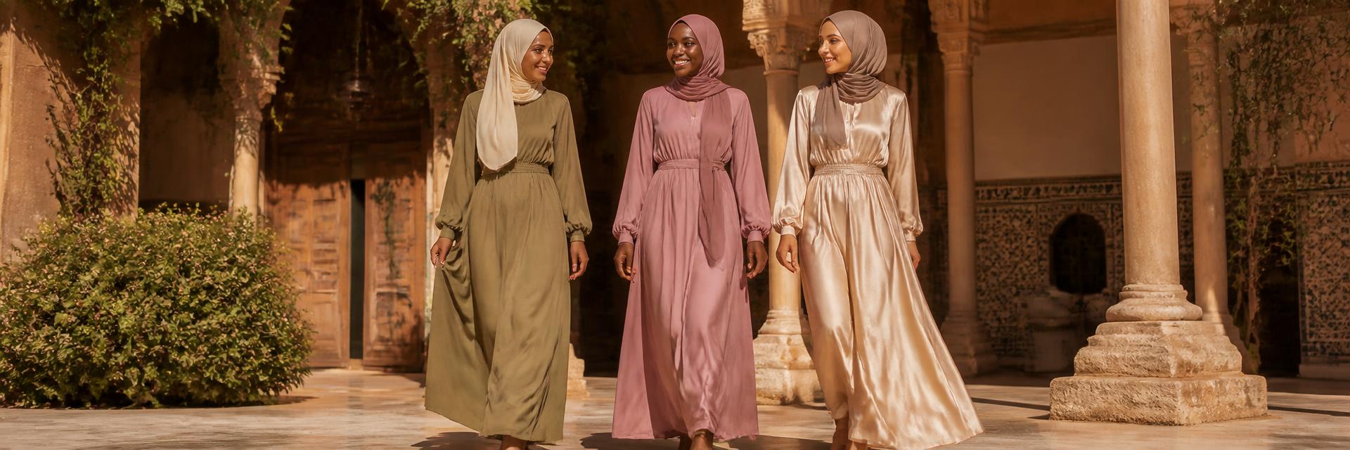 Three women — a Black woman center in a dusty rose maxi dress and matching hijab, a South Asian woman in olive green with cream hijab, a Middle Eastern woman in champagne satin with taupe hijab — walking through a sunlit Andalusian courtyard. Hair, neck and body fully covered.